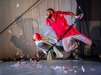 Vee Smith and Sadiq Ali hold onto a pole. They are gliding just above the floor and look as though they are flying. Both seem happy and content.