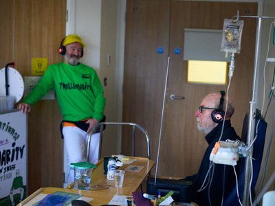 A man in a hospital bed has his eyes shut as he listens to the Trolleydarity story on his headphones. Alan looks on smiling.