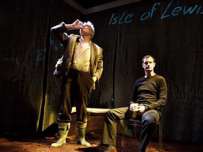 A man standing drinking from a glass, and beside him a man sitting in a chair