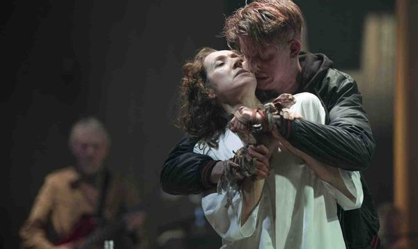 A distressed looking man dressed in dark clothes wraps his arms around a woman dressed in white. He holds on to her wrists and presses his face against her cheek.