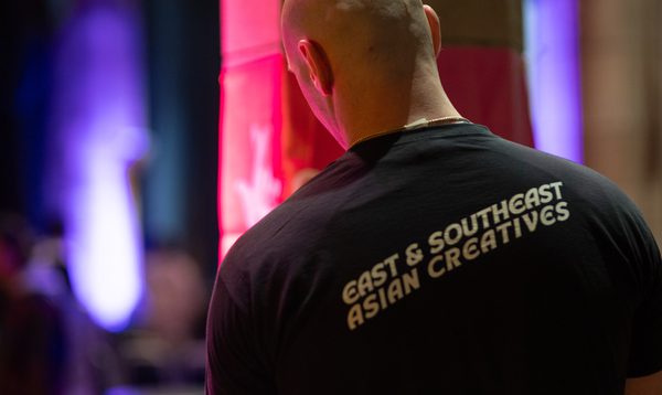 A bald crew member has his back to the camera. He is backstage and his t-shirt reads "East & South East Asian Creatives"