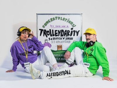 Two people are sat on either side of a poster for Trolleydarity. They are wearing branded tracksuits, one with a purple jumper and the other with a green jumper. They are both wearing yellow hats and headphones, one with a lanyard that says 'scratch' and the other that says 'sniff'.