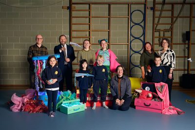 A group photo in a gym features adults and children with colorful props, including ribbons and suitcases. The mood is cheerful and creative, reflecting a playful atmosphere.