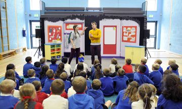 A group of school children in blue jumpers have their backs to the camera. They are looking towards to actors on a mobile set with has been erected in their school.