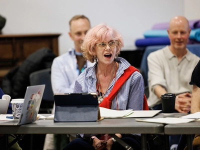 An actress with pink hair sits at a table her mouth is open in surprise two men sit behind her giggling.