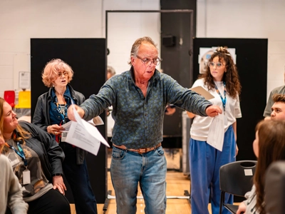 An older actor stand in the centre and he is gesturing with his arms, Other actors stand and sit around him watching.