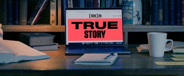 The room is dimly lit and a bookcase is in the background. There is a desk with a computer on it. The screen reads "True Story".