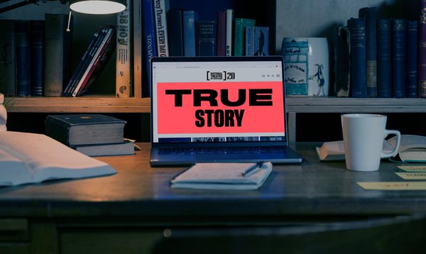 The room is dimly lit and a bookcase is in the background. There is a desk with a computer on it. The screen reads "True Story".