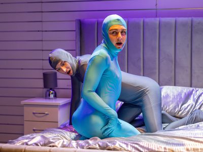 Two men wearing body suits sit on a bed, they look shocked.